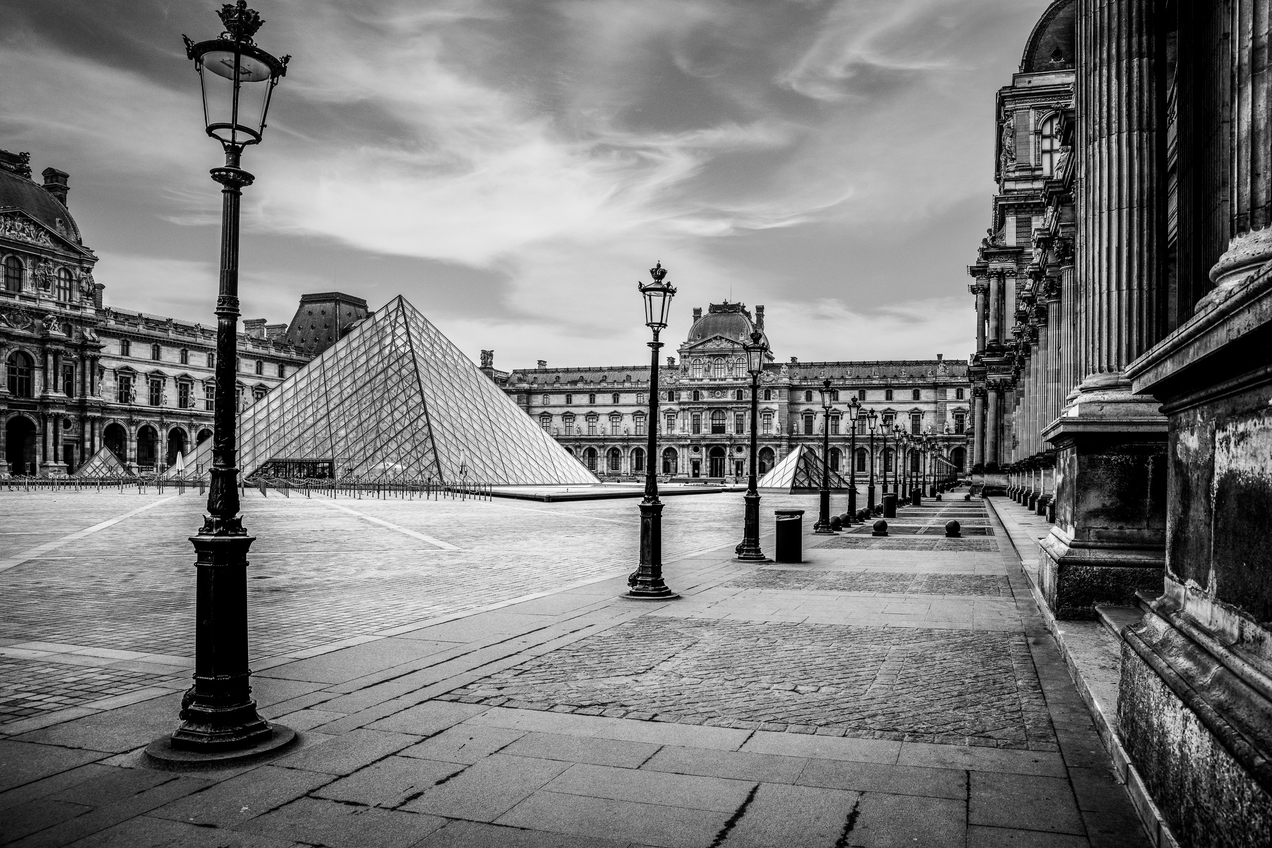 Horizontal black and white photograph of the Louvre Pyramid and historic courtyard in Paris with dramatic clouds and classical architecture