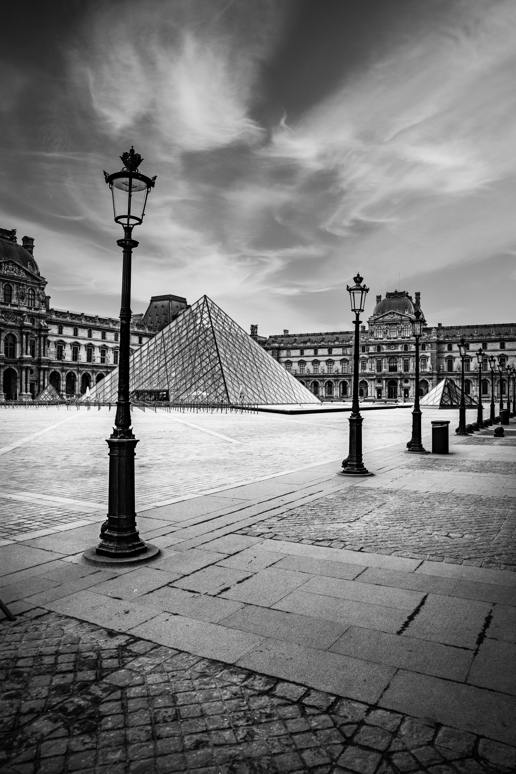 Black and white photograph of the Louvre Pyramid and courtyard in Paris with historic palace architecture and dramatic clouds