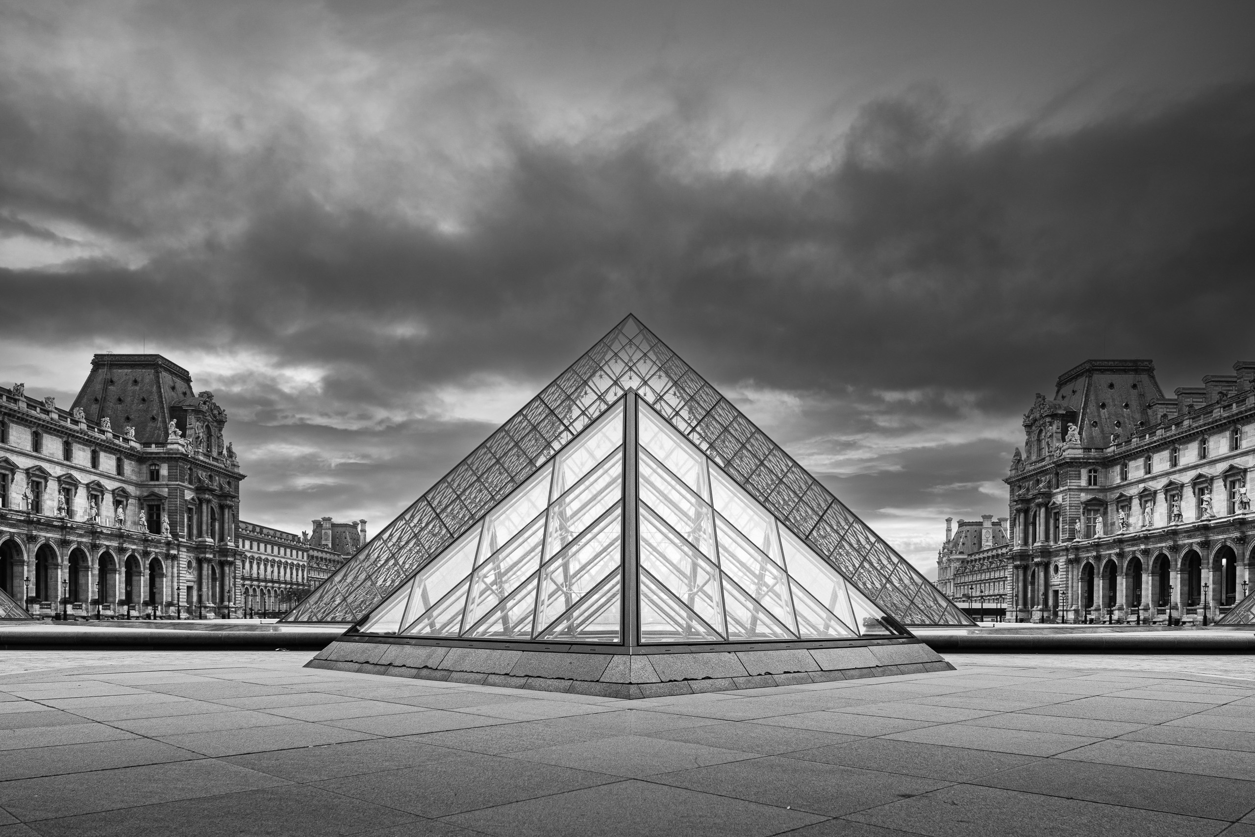Black and white photograph of the Louvre Pyramid in Paris, centered symmetrically beneath dramatic clouds in the historic courtyard.