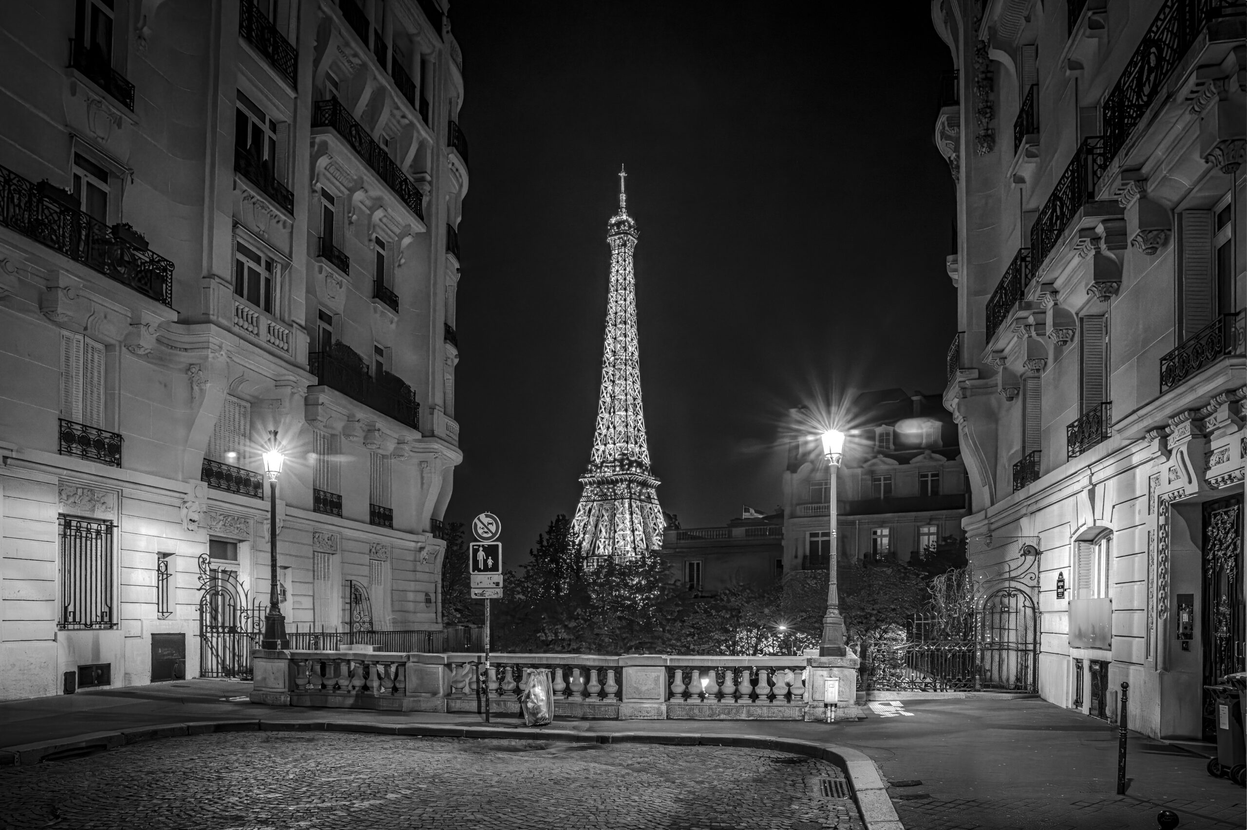 Black and white nighttime photograph of the Eiffel Tower illuminated between Parisian buildings