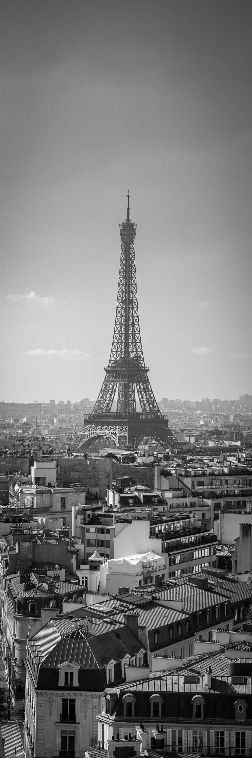 Vertical black and white panoramic photograph of the Eiffel Tower rising above Paris rooftops