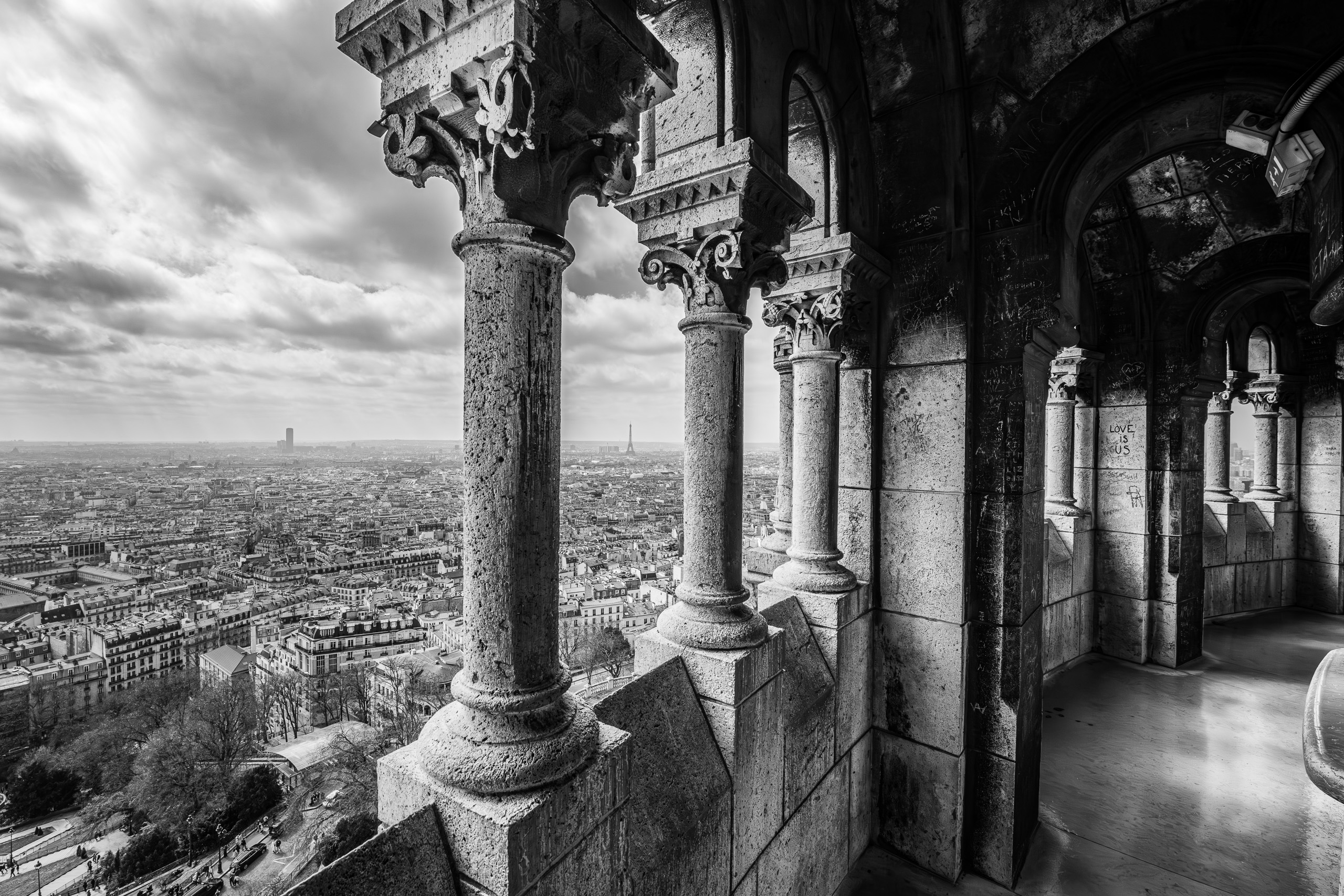 Black and white photograph of Paris viewed from the arches of Sacré-Cœur, with stone columns framing the distant Eiffel Tower.