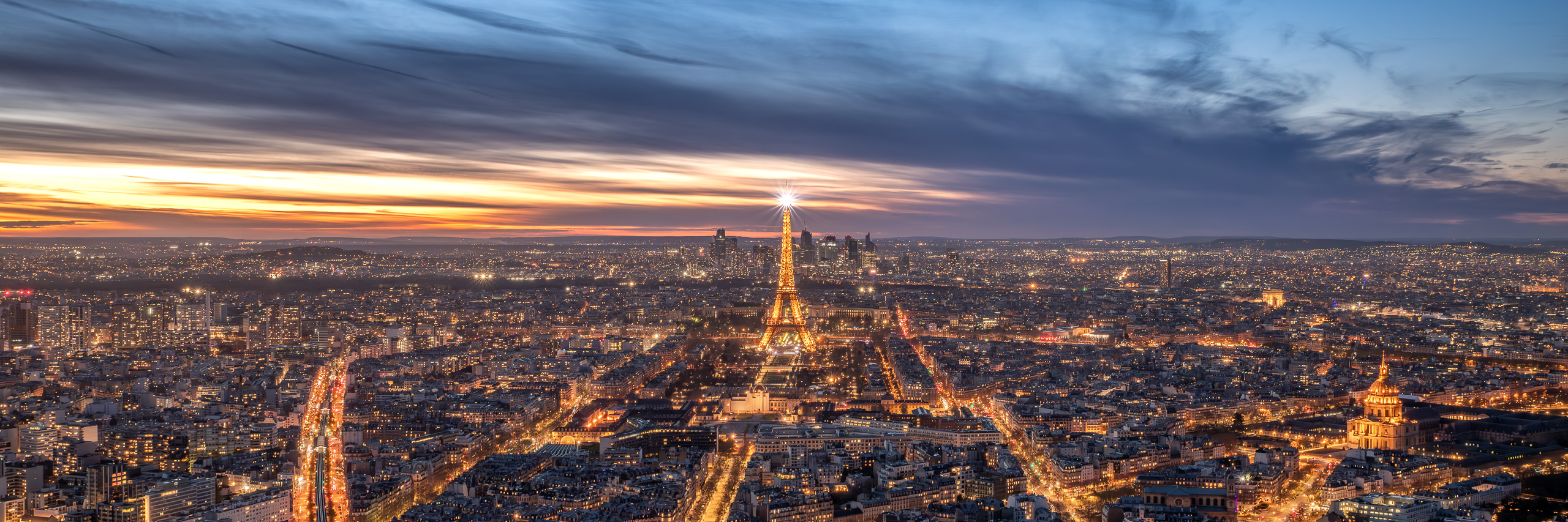 Panoramic night photograph of Paris with the Eiffel Tower illuminated and city lights stretching across the skyline.