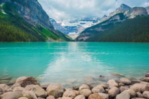Turquoise waters of Lake Louise with rocky shoreline, forest, and snowcapped mountains in Banff National Park Canada