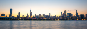 Panoramic sunset photograph of the Chicago skyline across Lake Michigan with city lights beginning to glow