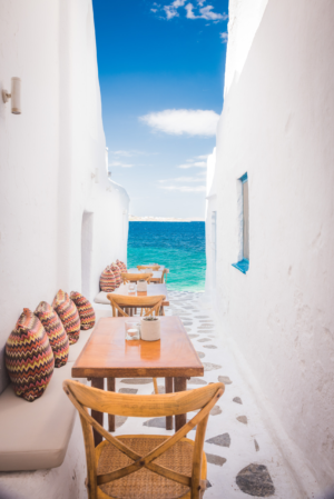 Narrow white alleyway with wooden tables and chairs overlooking turquoise sea in Mykonos Greece