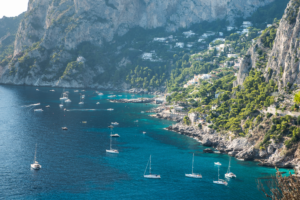 Horizontal photograph of Capri Italy coastline with turquoise ocean sailboats and dramatic cliffs