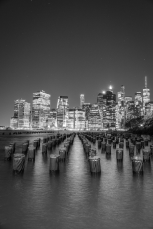 Black and white vertical photo of New York City skyline at night with old pier pilings in foreground
