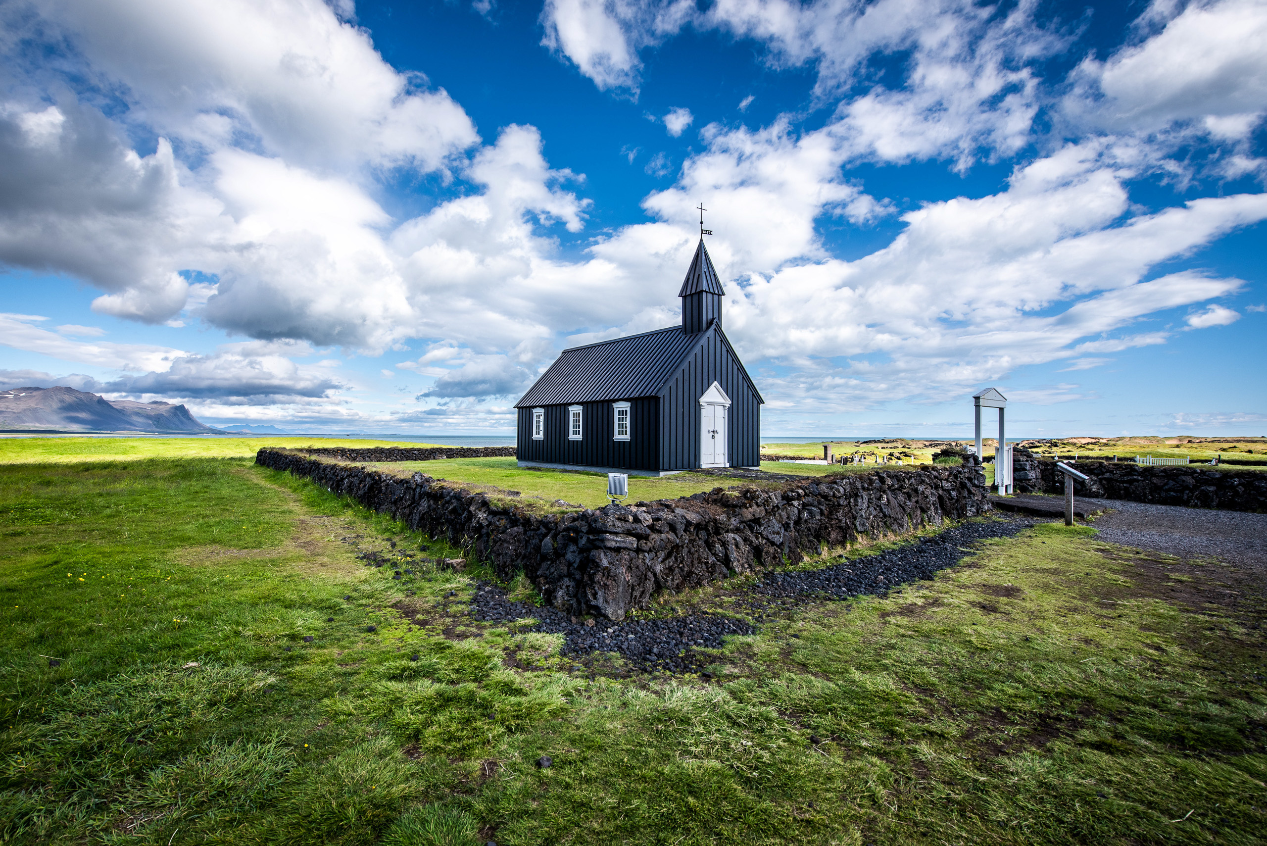 Color landscape photo of Budir Black Church in Iceland with blue sky clouds and lava stone wall