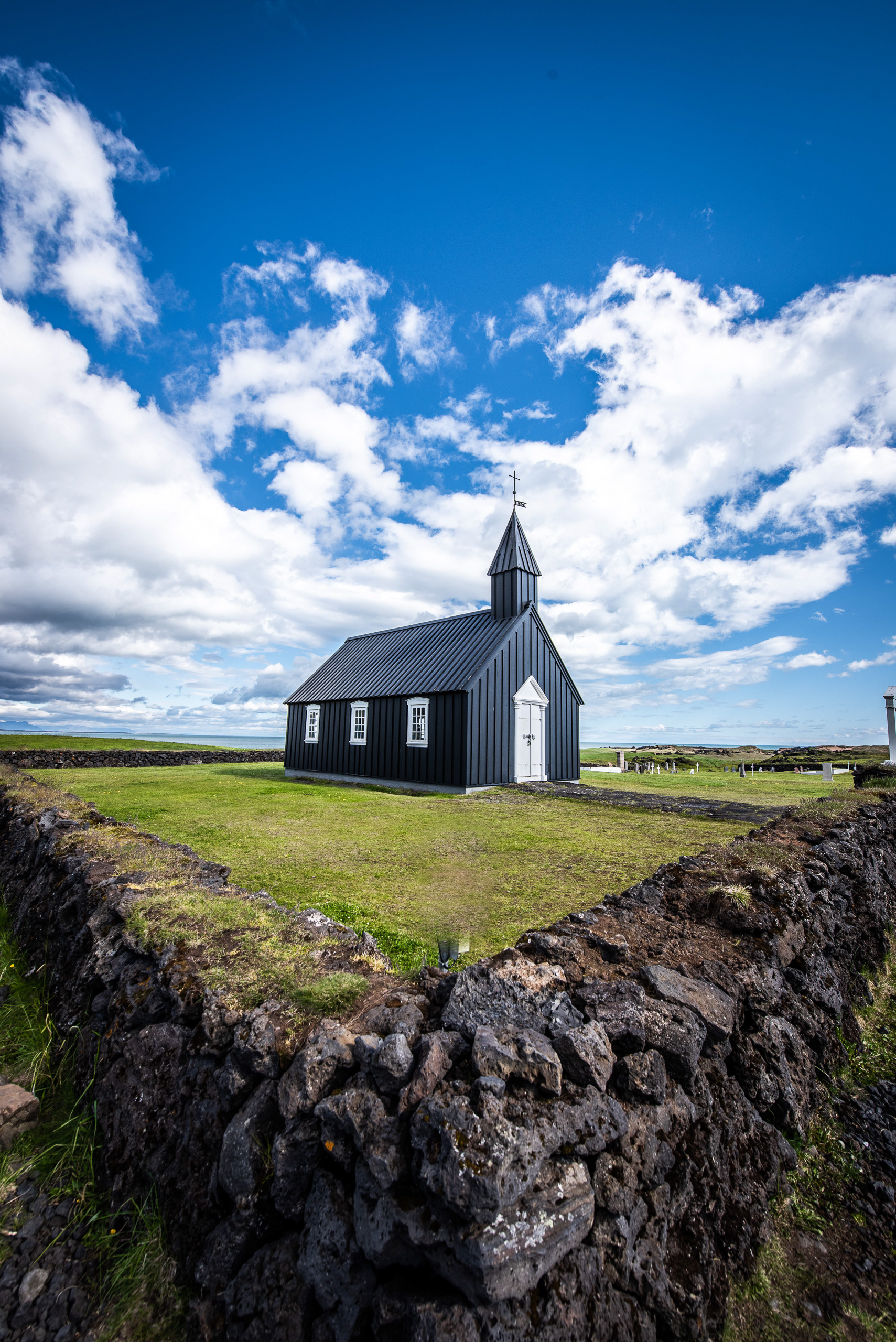 Vertical color photo of Budir Black Church in Iceland with blue sky clouds and lava rock wall