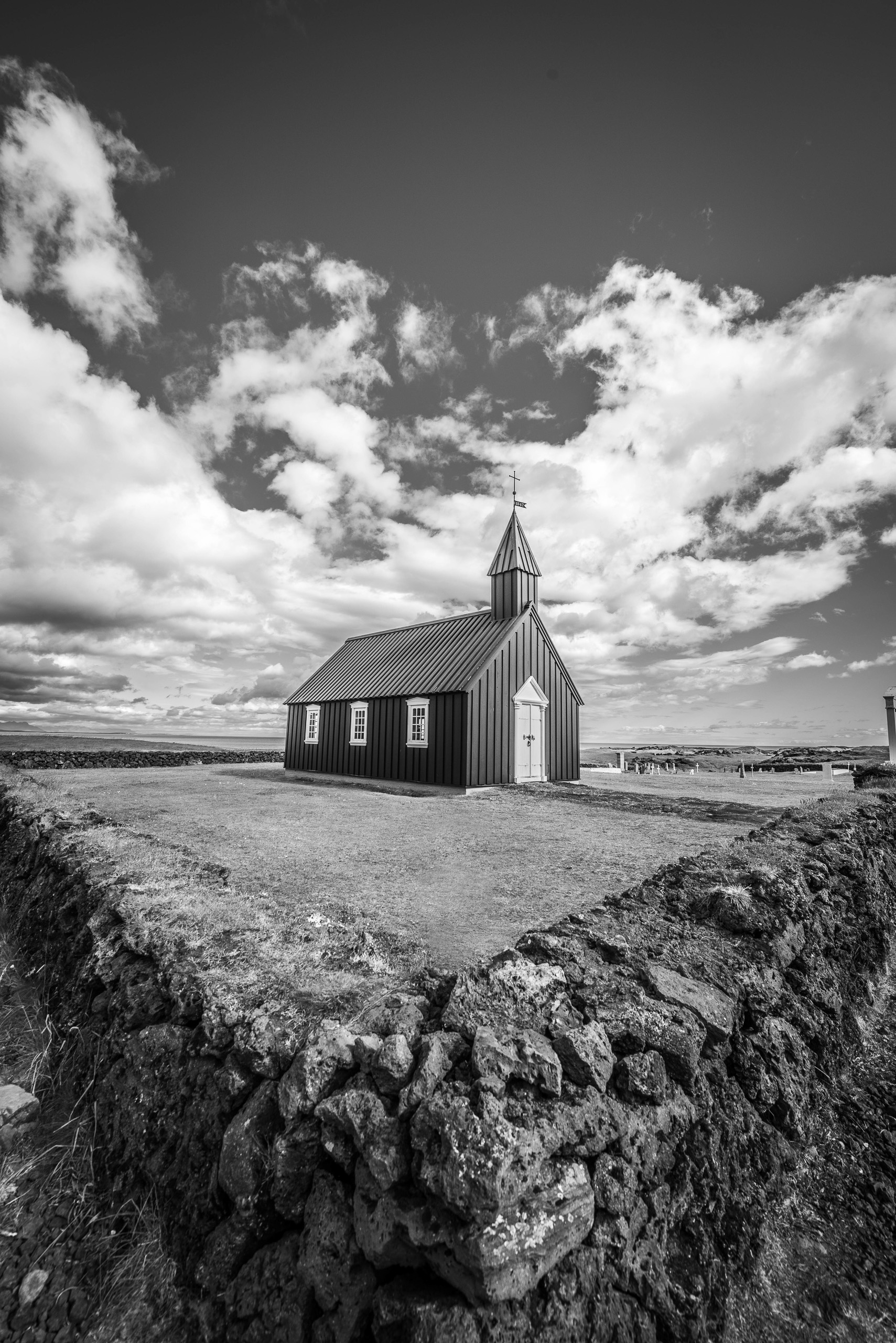 Vertical black and white photo of Budir Black Church in Iceland with clouds above lava stone wall
