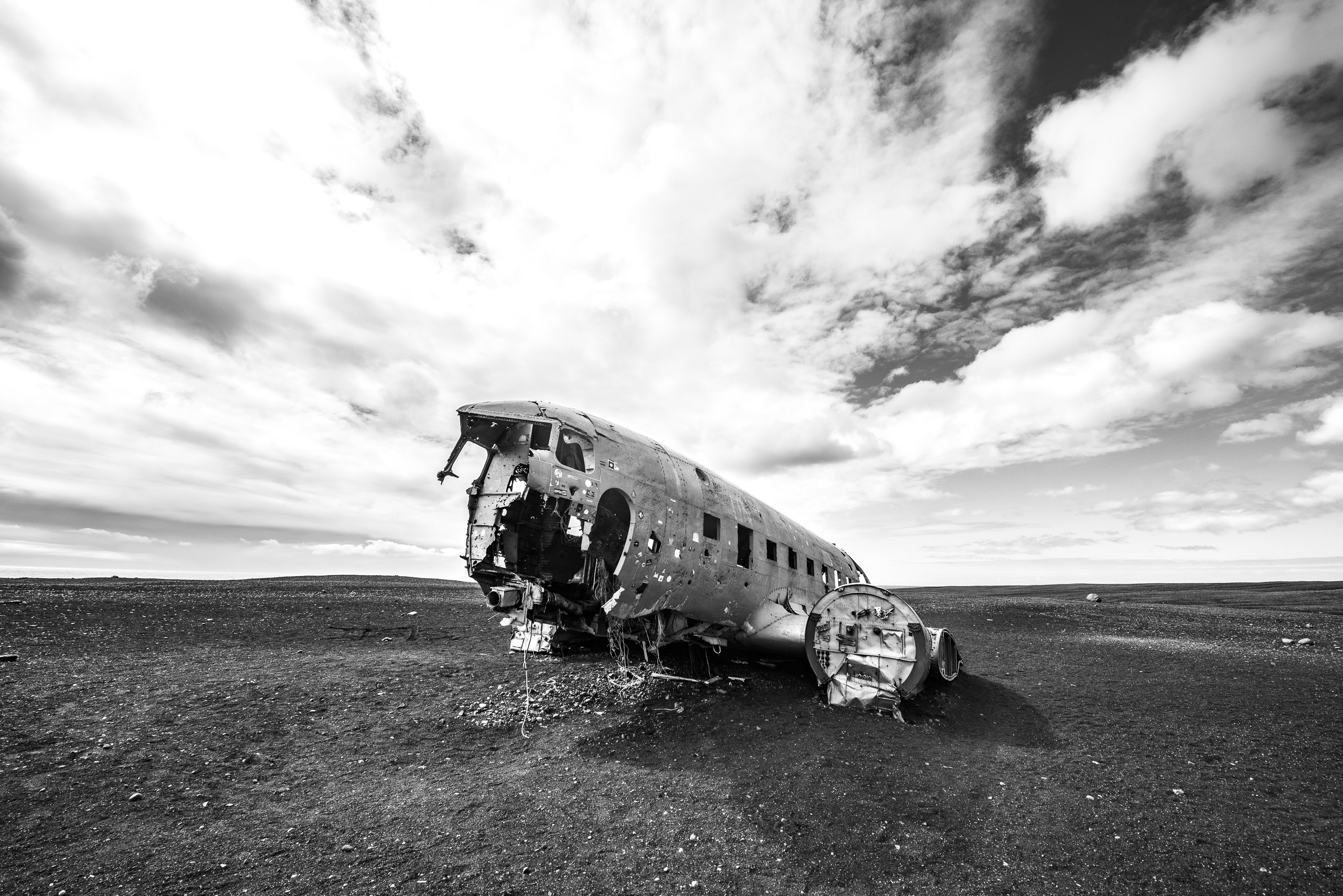 Black and white wide view of abandoned plane wreck on Iceland black sand plain beneath dramatic clouds