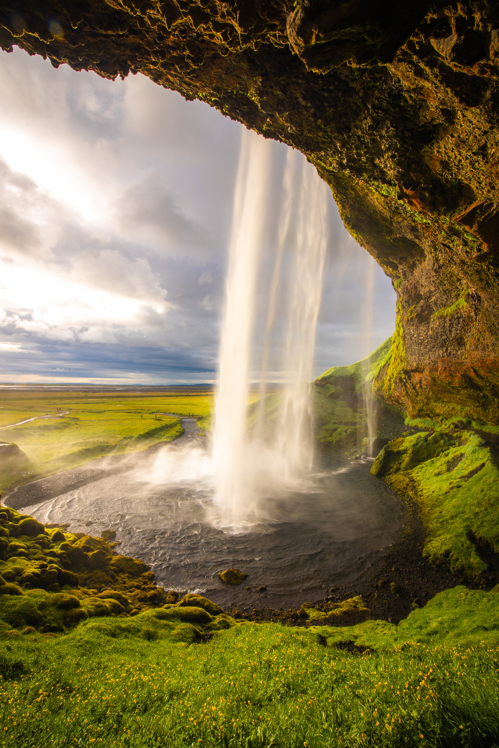 Vertical view of Seljalandsfoss waterfall in Iceland from behind with mossy cliffs and glowing sunlight