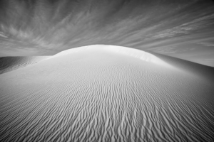 Black-and-white photograph of a towering gypsum dune at White Sands National Park, where intricate ripple patterns lead toward a brilliant dune crest beneath dramatic clouds.