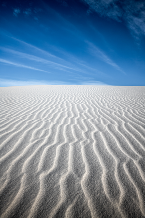 Brilliant white gypsum sand ripples stretch toward the horizon at White Sands National Park beneath a vivid blue sky streaked with delicate desert clouds.