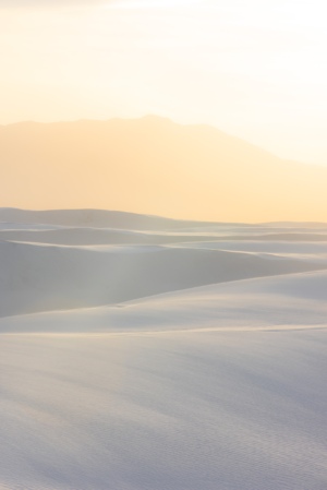 Soft sunrise light washes over the white gypsum dunes of White Sands National Park, with distant mountains glowing beneath a warm New Mexico sky.