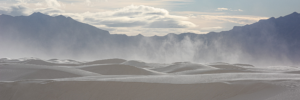 Windblown gypsum dunes stretch across White Sands National Park as drifting sand rises into the air beneath dramatic mountain peaks in southern New Mexico.