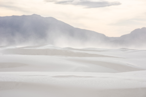 Windblown gypsum dunes stretch across White Sands National Park as drifting sand rises beneath rugged mountain peaks and a softly lit New Mexico sky.