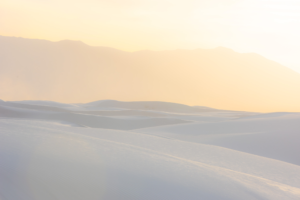 Warm sunrise light bathes the white gypsum dunes of White Sands National Park, creating soft layers of sand and pastel tones beneath distant New Mexico mountains.