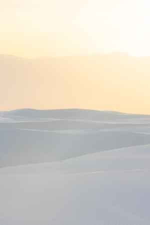 White gypsum dunes at White Sands National Park glow in soft sunrise light, with layered desert mountains fading into the distance beneath a warm New Mexico sky.